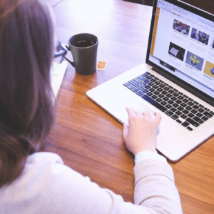 Person browsing a modern website on a laptop with a smartphone and coffee on a wooden desk.