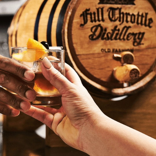 Two people clinking glasses of a cocktail in front of a Full Throttle Distillery whiskey barrel.
