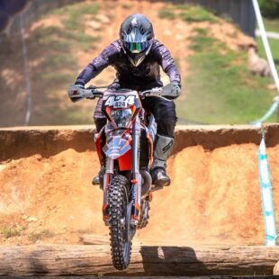 Sam on his dirt bike with the number 424, airborne after jumping a log obstacle under a Red Bull arch.