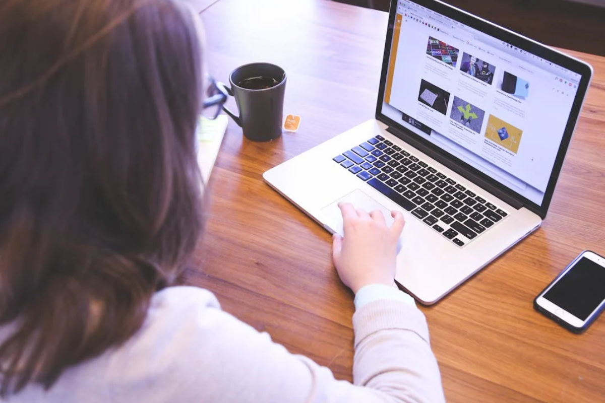 Person browsing a modern website on a laptop with a smartphone and coffee on a wooden desk.