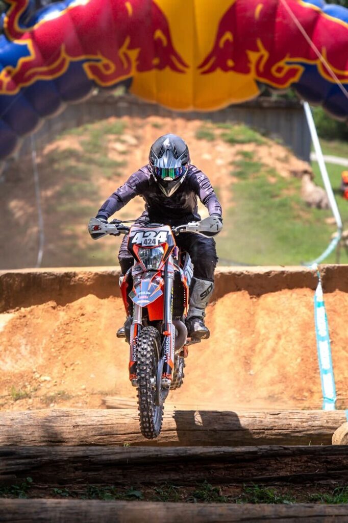 Sam on his dirt bike with the number 424, airborne after jumping a log obstacle under a Red Bull arch.