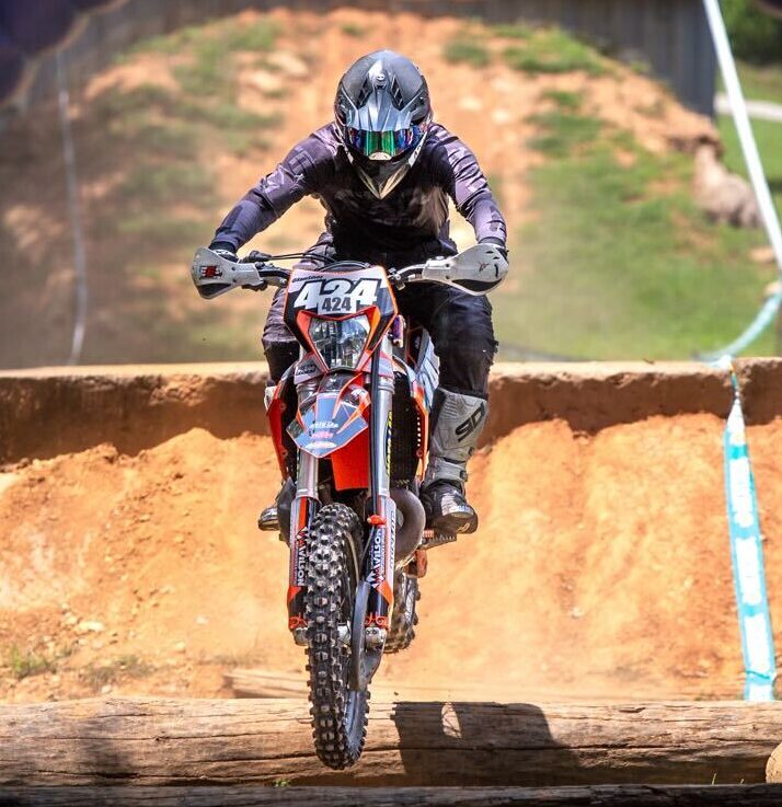 Sam on his dirt bike with the number 424, airborne after jumping a log obstacle under a Red Bull arch.