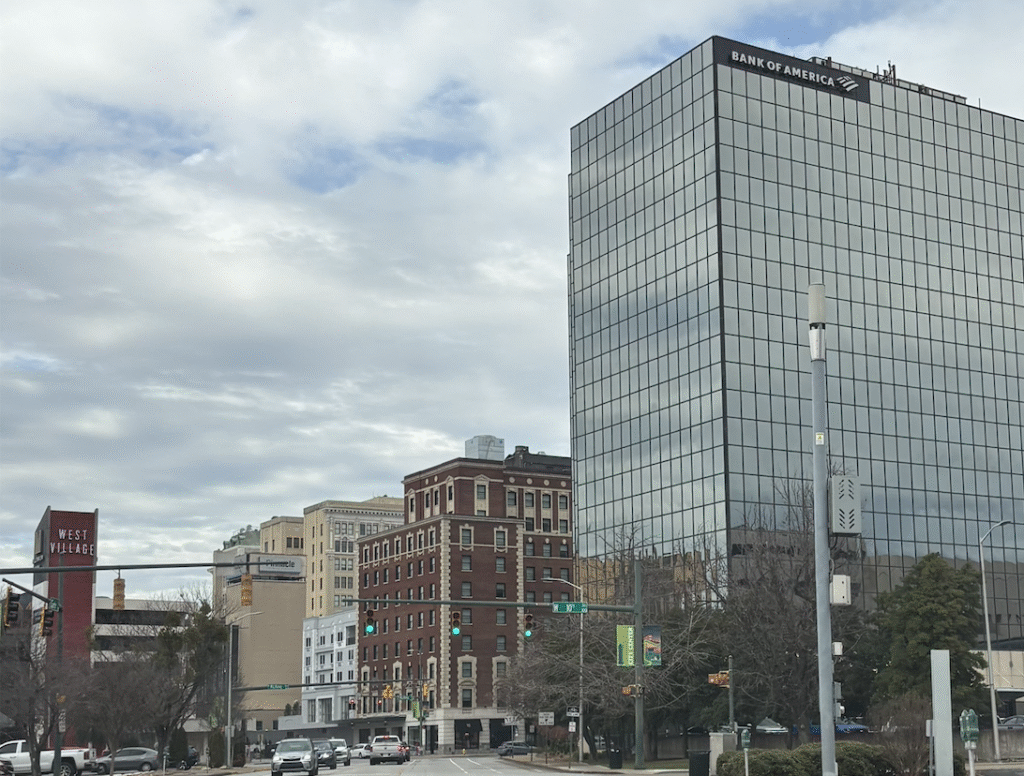 A view looking down West MLK Blvd in downtown Chattanooga under a cloudy sky. The sleek, glass-windowed Bank of America office building tower dominates the right side of the frame next to historic brick architecture and a street sign reading West 10th Street.