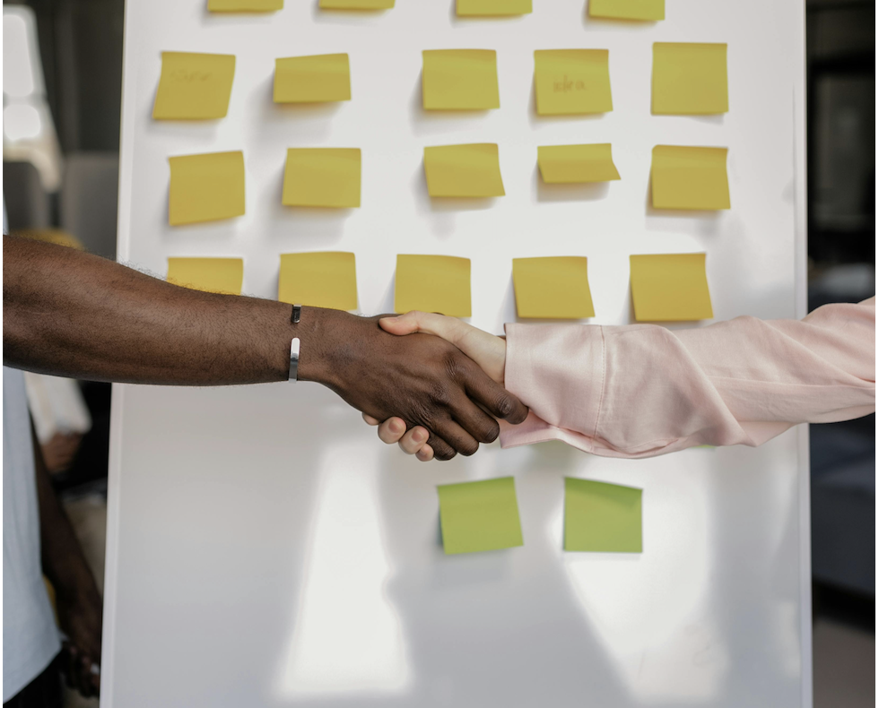 A close-up shot of a Black man and a woman shaking hands in a corporate setting. In the background, a white presentation board is covered in neat rows of yellow and green sticky notes, symbolizing strategy alignment and onboarding a marketing agency.