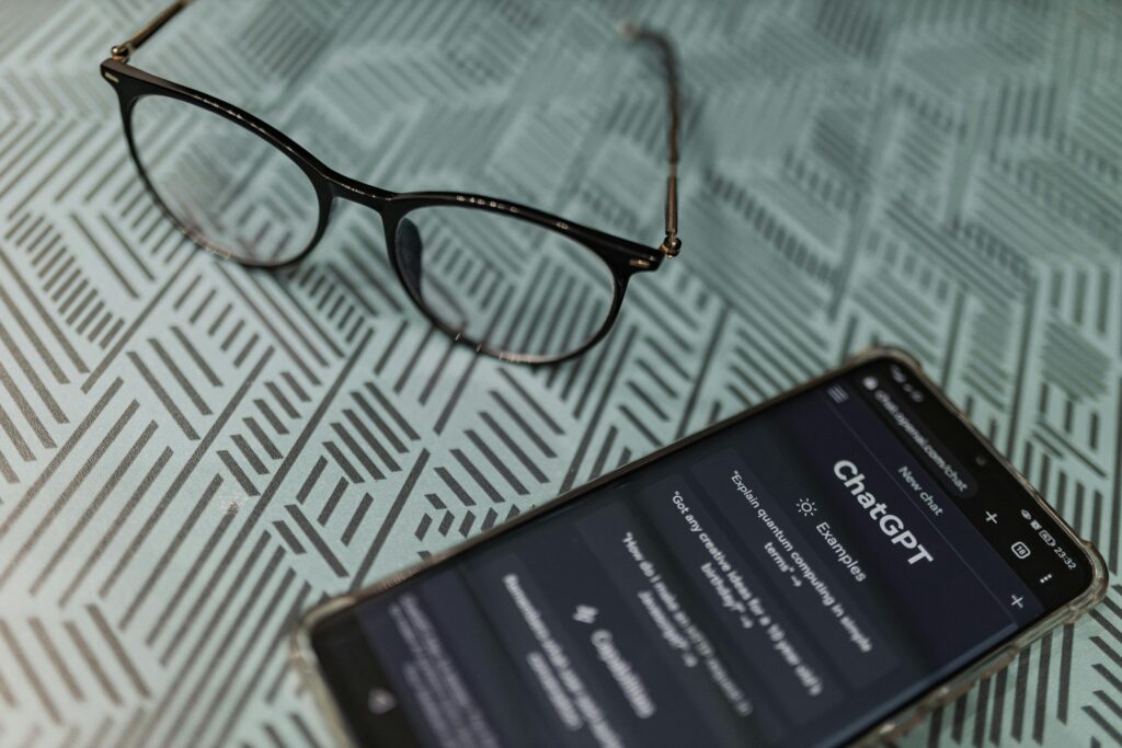 An overhead, angled shot of a smartphone displaying the ChatGPT home screen in dark mode with prompt examples like "Explain quantum computing in simple terms". A pair of black-rimmed eyeglasses sits next to the phone on a textured geometric pattern surface.
