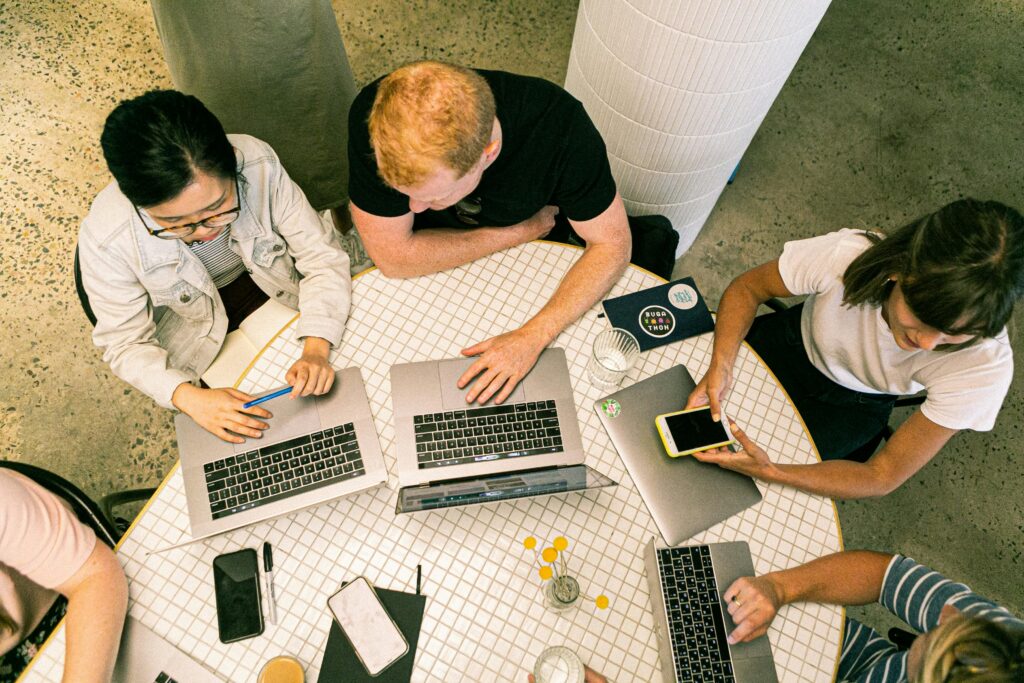 An overhead shot of a diverse group of five professionals at a marketing agency gathered around a white mosaic-tiled round table. They are actively collaborating using multiple open laptops, notebooks, and smartphones in a modern, well-lit workspace.