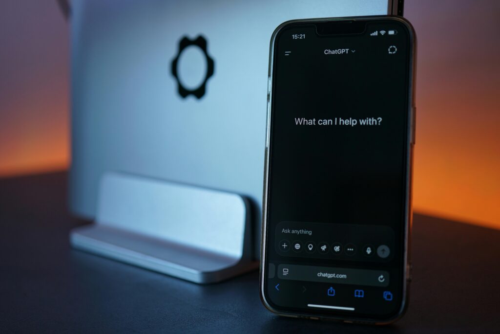 An iPhone displays the ChatGPT mobile search interface with the text "What can I help with?" next to a vertically docked Framework laptop. This highlights Generative Engine Optimization (GEO) and the shift from traditional search engines to AI assistants.