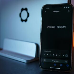 An iPhone displays the ChatGPT mobile search interface with the text "What can I help with?" next to a vertically docked Framework laptop. This highlights Generative Engine Optimization (GEO) and the shift from traditional search engines to AI assistants.