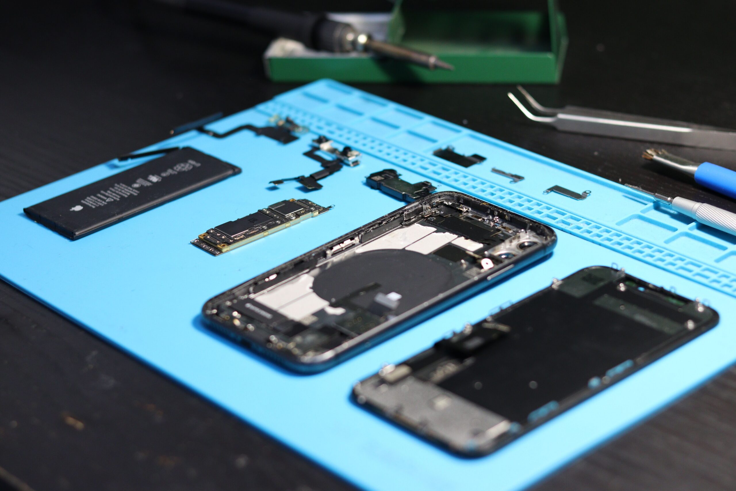 Disassembled iPhone on a blue repair mat with a soldering iron and tweezers, representing the Right to Repair movement